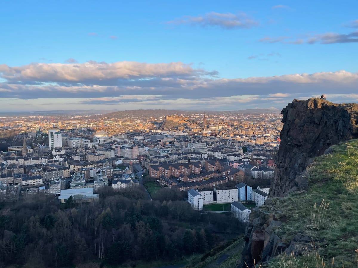 A city skyline at sunrise, viewed from above with grassy-rocky hill in the foreground. 