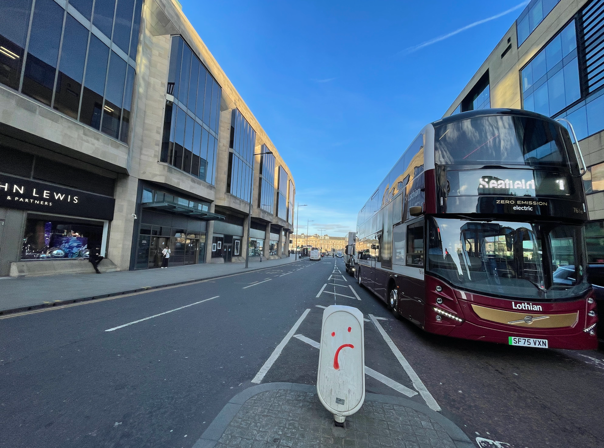 A bus on a city street with tall buildings on either side of the road below a blue sky. 