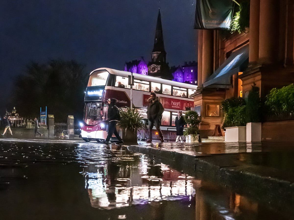 People walking in the rain through a city centre past a bus next to a hotel, with a castle in the background. 