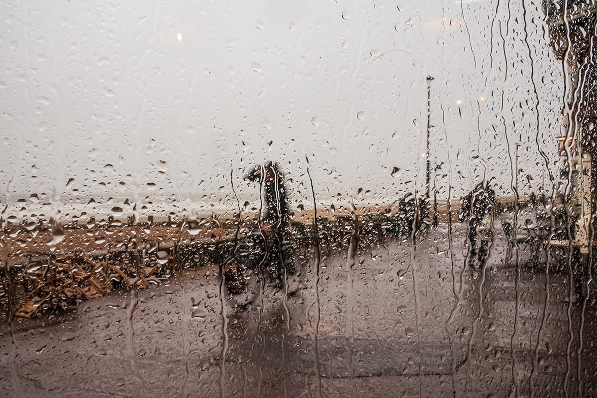 A person cycling on a beachside path, seen through a rain-covered window. 