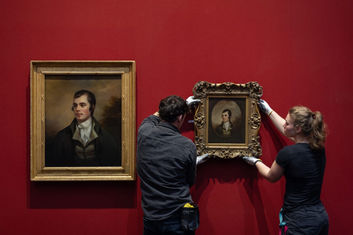 Two people hanging a framed painting on a red wall next to a similar but larger portrait. 