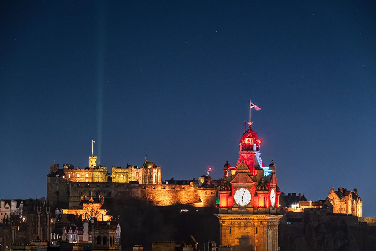 A feint yellow beam of light in the night sky above Edinburgh Castle, with a clock tower in the foreground. 