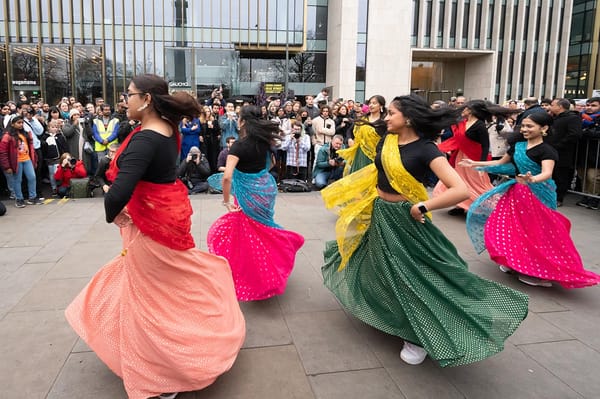 People wearing colourful outfits dancing in an outdoor paved public square surrounded by a crowd of people.