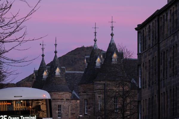 Silhouettes of building roofs against a pink sky with a hill in the background and a bus in the foreground. 