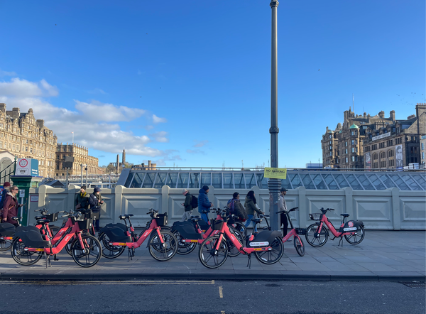 People walking past a group of red hire bikes on a bridge in a city centre under a blue sky. 
