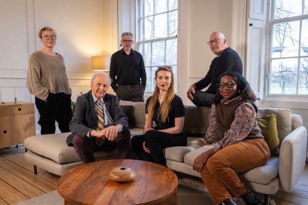 Six people, three standing and three sitting on a sofa, in a high-ceilinged living room with two large windows, wooden flooring, plus a circular wooden table in the foreground. 