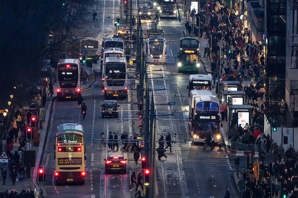 Four lanes of traffic flanked either side by pedestrians on a city street.