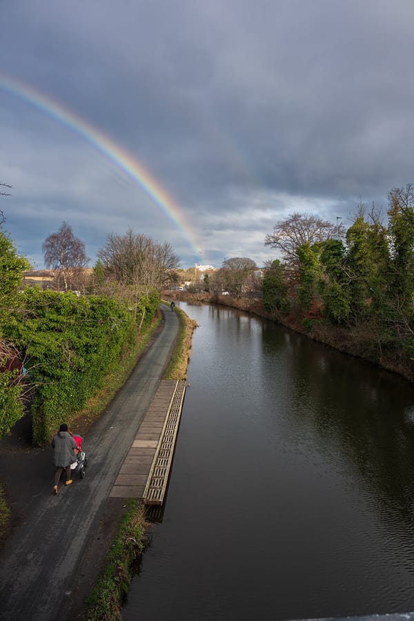 A rainbow over a canal, with a person pushing a buggy in the foreground. 